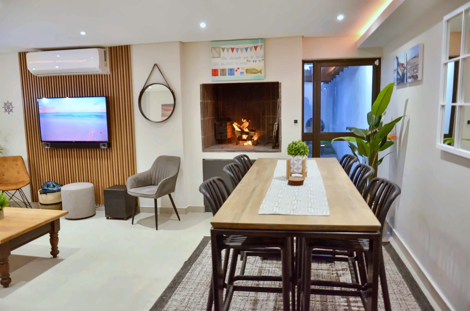 Open-plan living and dining area with the braai fire lit and a TV on a slat wall